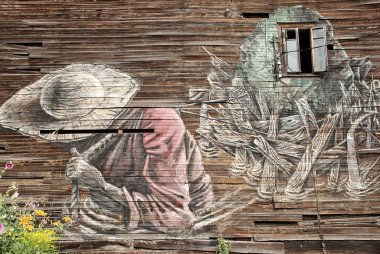Bir evin ahşap duvarına grafiti. Hasır şapkalı ve kırmızı gömlekli bir adam. Taşı iple sürüklüyor.