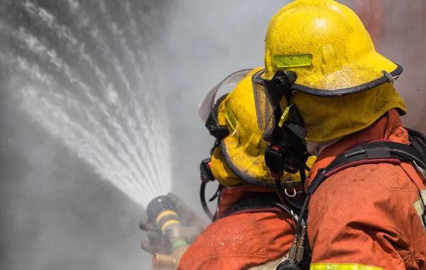 Due pompieri in casco e maschera di ossigeno spruzzando acqua a fuoco su — Foto Stock