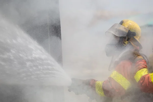 Bombero en traje de extinción de incendios rociando agua para fuego envolvente wi —  Fotos de Stock