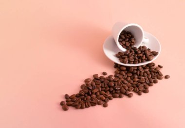 Açık pembe arka planda kahve çekirdekleri dökülmüş bir fincan kahve.