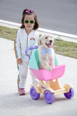 Beyaz elbiseli ve güneş gözlüklü güzel çocuk Golden Retriever köpeğiyle oynuyor. .