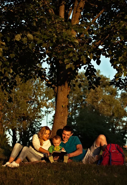 Familia feliz en el parque relajándose juntos y leyendo el libro para el pequeño hijo . —  Fotos de Stock