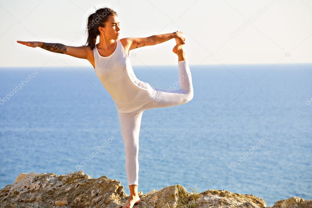 Slim athletic young girl doing yoga on the rock face on the background of blue sea.