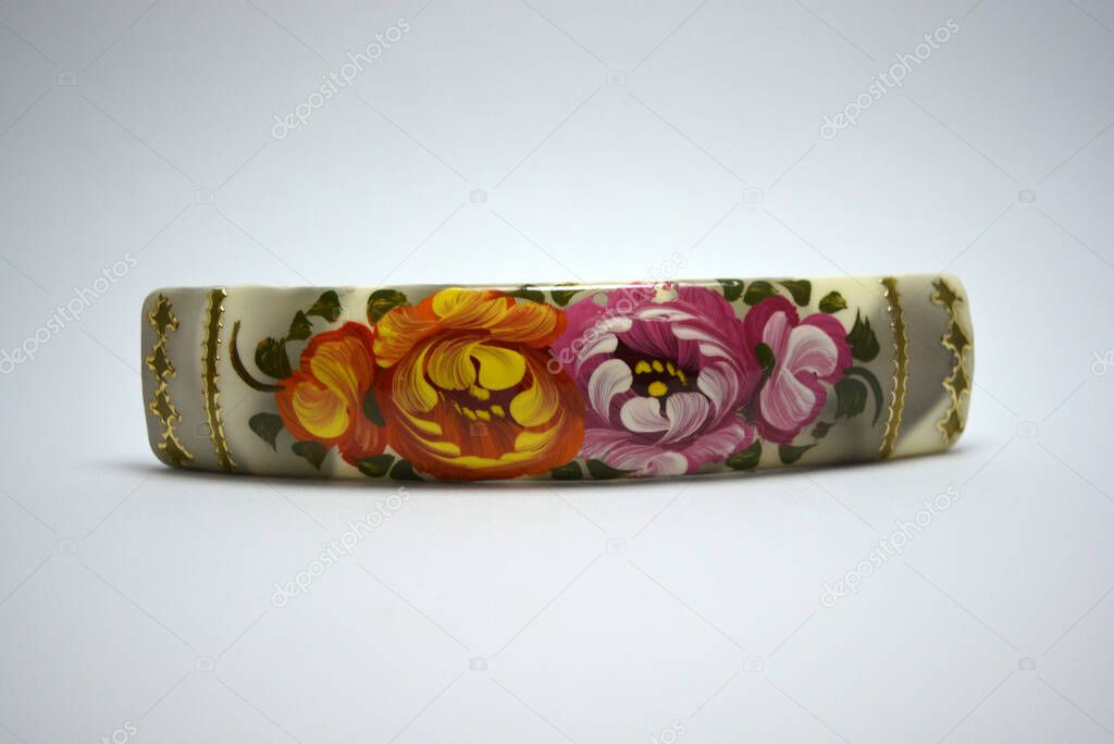 A beautiful and stylish metal hair clip decorated with the national Petrikov painting in the form of original and unusual bright colors on a white wooden background. 