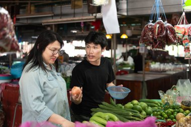 İki LGBTQ arkadaşı kalabalık bir Tayland pazarından taze sebze alıyor. Biri sebzeleri seçerken, diğeri ilgiyle bakıyor, dostça bağlarını canlı, günlük bir sahnede sergiliyor..