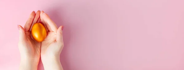 Golden Easter egg in hand on a pink background. Happy spring holiday. Banner format