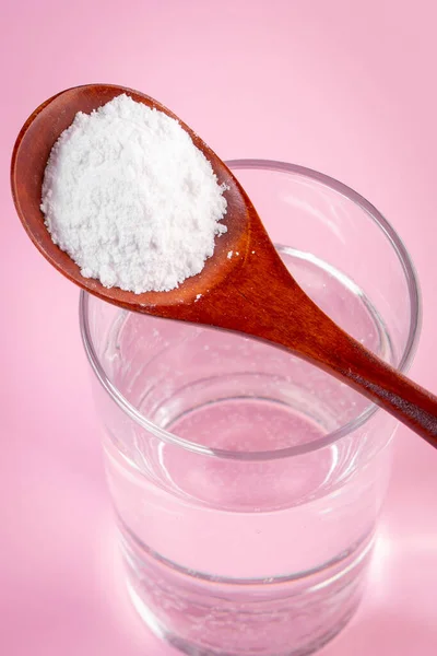 Natural bio supplement collagen powder in wooden spoon and glass of pure water on pink background. Concept healthy skin and bones, anti-aging extra protein product. Flatlay, place for text.