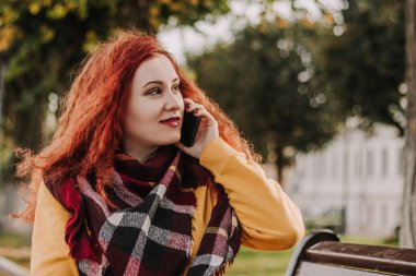 Sarı kazaklı, genç, kızıl saçlı kadın telefonda konuşuyor. Bayan sonbahar günü bankta oturur. Yaşam biçimi.