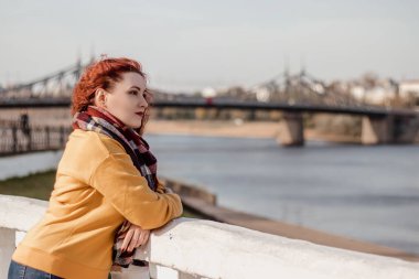 Genç kızıl saçlı kadın sonbahar güneşli bir günde rıhtımda yürüyor ve manzaranın tadını çıkarıyor. Bayan mesafeye bakıyor..