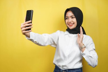 Güzel, genç Asyalı Müslüman kadın kendinden emin, coşkulu ve neşeli, akıllı telefonu tutan, birini arayan, video görüşmesi yapan, selfie çeken, sarı arka planda izole edilmiş, reklamcılık konsepti olan