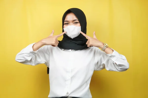 Muslim woman wearing white mask, with hand pointing to mask, prevent corona virus gesture, prevent covid-19, isolated on yellow background