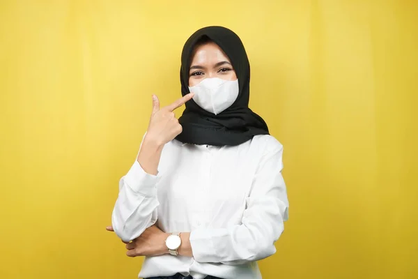 Muslim woman wearing white mask, with hand pointing to mask, prevent corona virus gesture, prevent covid-19, isolated on yellow background