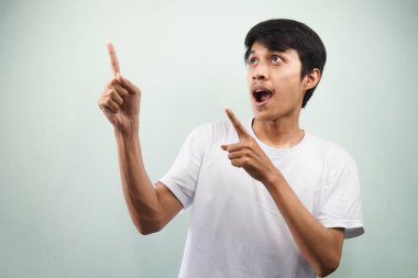 Beyaz tişörtlü genç Asyalı bir adam iki eliyle gri arka planda izole edilmiş boş bir yeri işaret ederek şaşırdı.