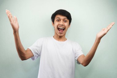 Beyaz tişörtlü genç Asyalı adam heyecanlı ve kendinden emin bir şekilde kameraya bakıyor. İki elini de kaldırmış gri arka planda tek başına.