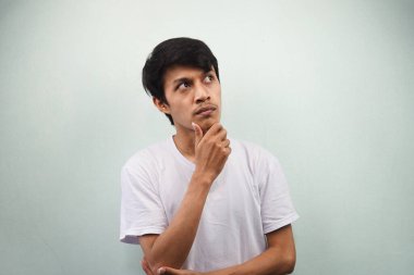 Beyaz tişört giyen Asyalı genç adam kafası karışmış ve gri arka planda izole edilmiş boş bir yere bakıyor.