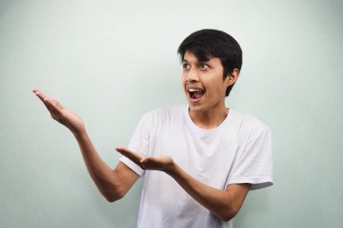 Beyaz tişörtlü genç Asyalı adam heyecanlı ve güvenli bir şekilde boş alana bakıyor. İki eli de gri arka planda, reklamcılık kavramında izole edilmiş.