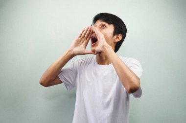 Beyaz tişörtlü genç Asyalı bağırıyor ve gri arka planda izole edilmiş boş alana doğru bakıyor.