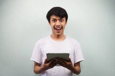 Beyaz tişörtlü genç Asyalı elinde tablet, gülümseyen mutlu ve kendine güvenen gri arka planda izole edilmiş kameraya bakıyor.