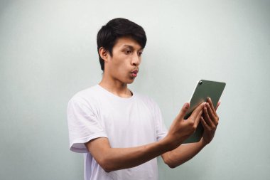 Beyaz tişörtlü, elinde tablet olan, gri arka planda, reklam konseptine izole edilmiş, şaşırmış bir ifade taşıyan Asyalı genç bir adam.