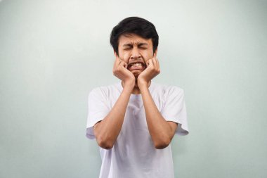 Beyaz tişört giyen Asyalı genç adam strese girmiş, paniklemiş, el sıkışıyor gri arka planda yalnız başına kafa tutuyor.