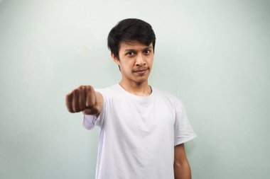 Beyaz tişörtlü genç Asyalı adam gri arka planda izole edilmiş kameraya yumruğunu sıkıp güvenle gülümsüyor.