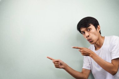 Beyaz tişörtlü genç Asyalı bir adam iki eliyle gri arka planda izole edilmiş boş bir yeri işaret ederek şaşırdı.