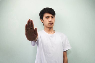 Beyaz tişört giyen Asyalı genç adam. Elinde dur işareti olan ciddi bir ifade var. İçki yasağı, gerileme, gri arka planda izole edilmiş kameraya doğru elini aç.