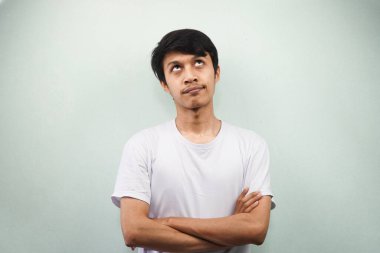 Beyaz t-shirt giyen genç Asyalı bir adam bir fikir edinmek istiyor. Gri arka planda izole edilmiş bir yere bakıyor.