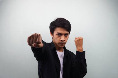 Siyah gömlekli genç Asyalı ciddi ve güçlü bir dövüş sanatları ustası, yumruğunu sıkıyor, kameraya yumruk atıyor, dövüşüyor, ruhuna hakim oluyor ve gri arka planda dövüşmeye hazır.