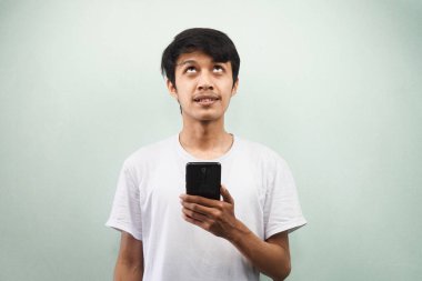 Beyaz tişörtlü genç Asyalı adam gülümsüyor, boş yere bakıyor, elinde akıllı telefon, gri arka planda izole edilmiş, reklam konsepti.