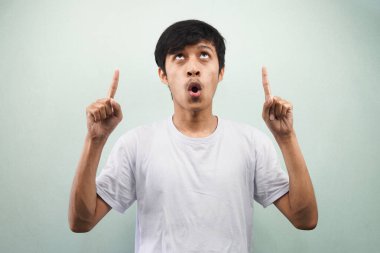 Beyaz tişört giyen Asyalı genç adam şaşırdı. Elleri havada, ağzı açık, gözleri açık gri arka planda izole edilmiş boş bir yere bakıyordu.