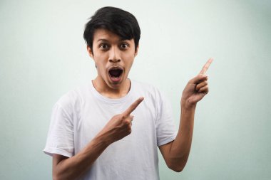 Beyaz t-shirt giyen genç Asyalı adam şaşırdı, vay be yüz ifadesi, eli boş alanı işaret ederek bir şey sunuyor, gri arka planda izole kameraya bakıyor, reklam kavramı