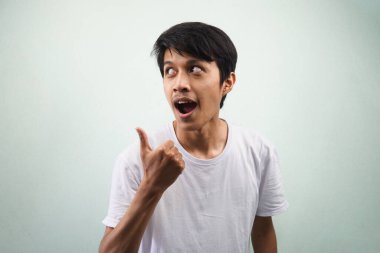 Beyaz tişört giyen Asyalı genç adam heyecanla ve kendinden emin bir şekilde gülümsüyor. Gözleri boş yere bakıyor ve başparmakları gri arka planda izole edilmiş bir şey sunuyor. Reklam konsepti.