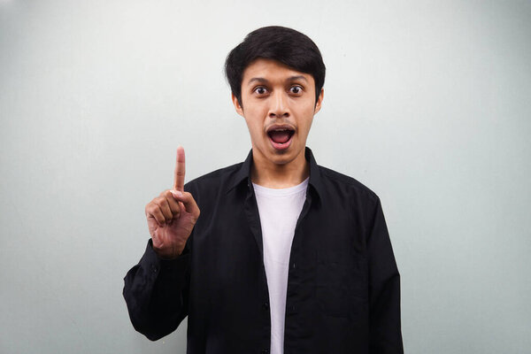 Молодой азиат, одетый в черную рубашку, удивился, получил идею, вспомнил что-то, с открытым ртом и пальцами, указывающими на изолированный на сером фоне
