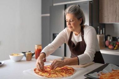 Üst düzey bir kadın önlük giyiyor, modern bir mutfakta ev yapımı pizza yapıyor.
