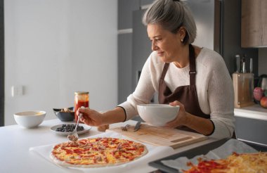 Son sınıf öğrencisi kadın pizza hazırlıyor, mutfakta hamura malzeme ekliyor.