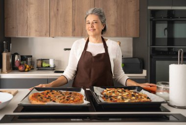 Son sınıf kadını gülümsüyor, mutfakta elinde fırında pişmiş iki pizzayla duruyor.