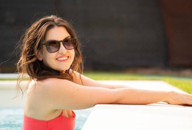 Güneş gözlüklü gülümseyen kadın yüzme havuzunda yaz tatilinin tadını çıkarıyor.