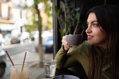 Kahve içen bir kadın, gülümseyen ve kafenin penceresinden dışarı bakan, rahatlamış hisseden.