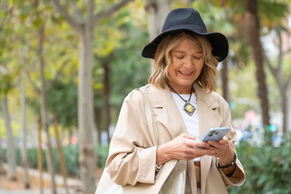 Şehir parkında akıllı telefon kullanan gülümseyen son sınıf öğrencisi kadın, kopyalama alanı teknolojisiyle bağlantılı.