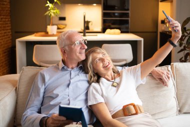 Mutlu son sınıf çifti kanepede dinleniyor, modern evde akıllı telefonuyla selfie çekiyor.