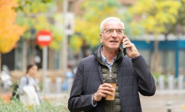 Kıdemli adamın ön görüntüsü bir telefon görüşmesi yapıyor, elinde bir kahve fincanı tutuyor ve elinde fotokopi aletiyle dışarı çıkıyor.