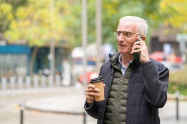 Kıdemli adam gülümsüyor, telefonda konuşurken dışarıda yürüyor ve elinde fotokopi alanı olan bir kahve fincanı taşıyor.