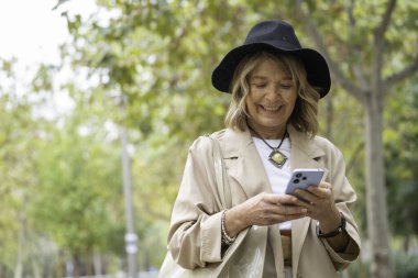 Kıdemli kadın gülümsüyor, cep telefonuyla dışarıda fotokopi çektiriyor.