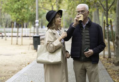 Yaşlı çift parkta rahatça yürürken sohbet ve kahveden hoşlanıyor.