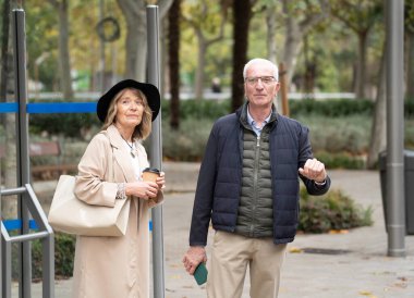 Şehir parkında otobüs durağında bekleyen yaşlı çift, toplu taşıma için hazırlar.