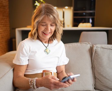 Mutlu olgun bir kadın evde oturup telefonuyla sosyal medyayı karıştırıyor.