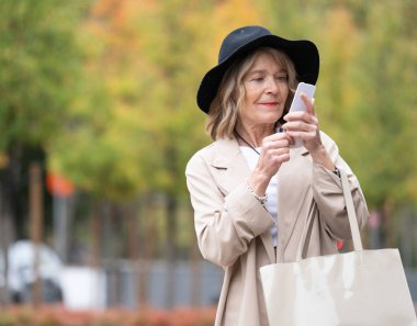 Trençkotlu ve şapkalı yaşlı bir kadın açık havada fotokopi alanıyla iletişim kuruyor.