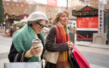 İki son sınıf bayan arkadaş açık hava Noel pazarında kahve ve alışveriş torbalarıyla eğleniyorlar.