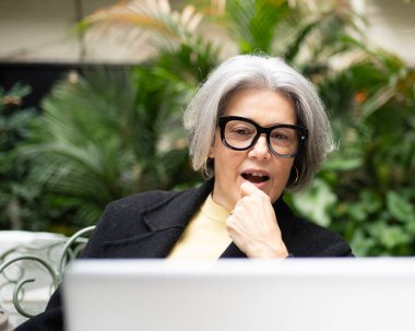 Beyaz saçlı bir kadın dışarıda çalışırken laptoptan gelen bilgilere tepki veriyor.